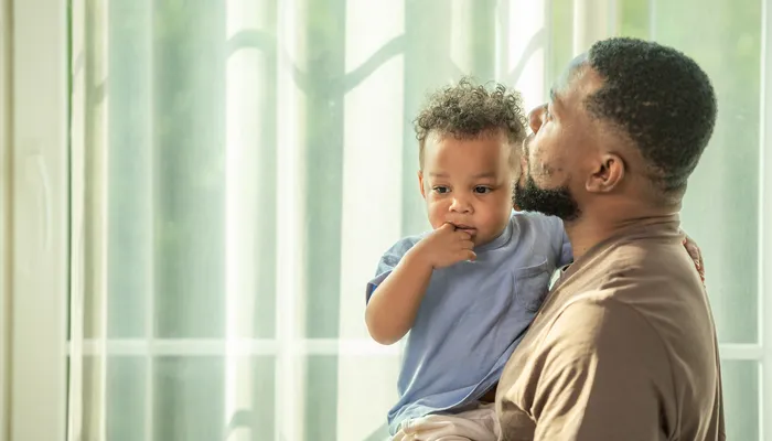 An adult male holding a young child indoors near a window with sheer curtains, both facing sideways. The scene conveys a tender moment with soft natural light highlighting their close interaction.