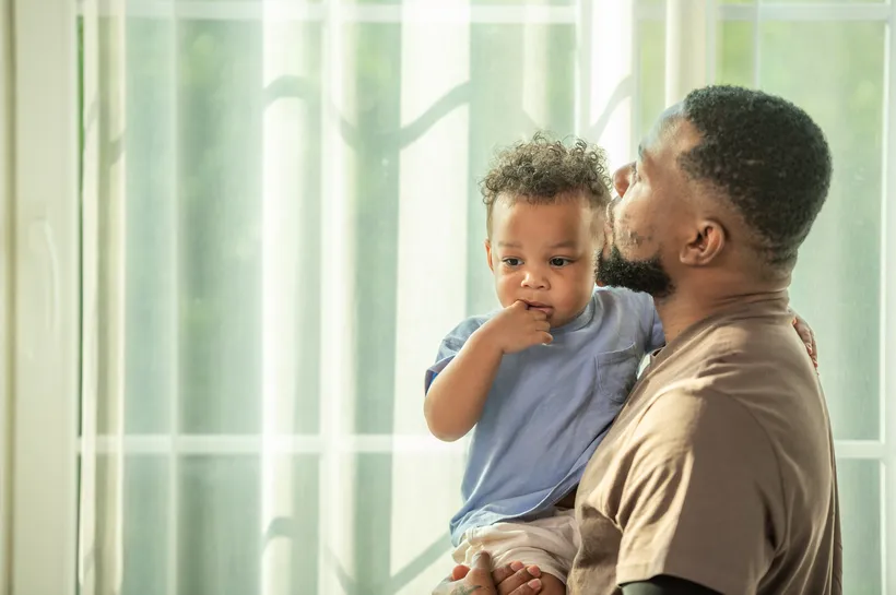 An adult male holding a young child indoors near a window with sheer curtains, both facing sideways. The scene conveys a tender moment with soft natural light highlighting their close interaction.