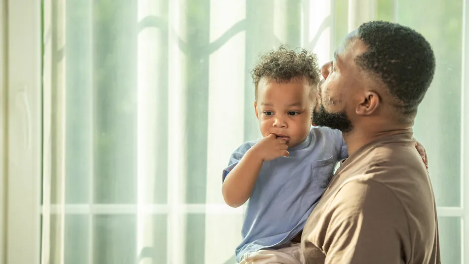 An adult male holding a young child indoors near a window with sheer curtains, both facing sideways. The scene conveys a tender moment with soft natural light highlighting their close interaction.