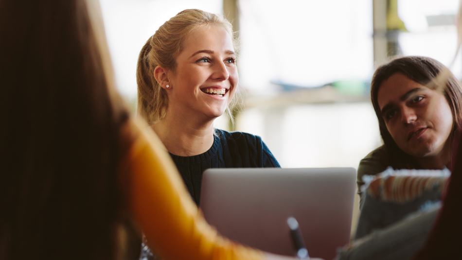 A smiling young person sitting with three people around a table, one using a laptop. The setting appears to be a casual or collaborative workspace with natural light in the background.