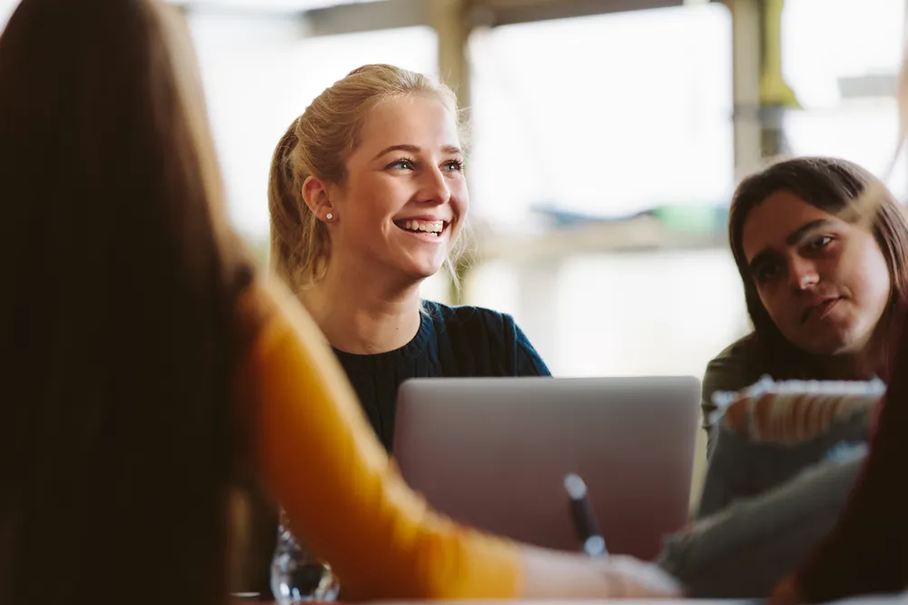 A smiling young person sitting with three people around a table, one using a laptop. The setting appears to be a casual or collaborative workspace with natural light in the background.