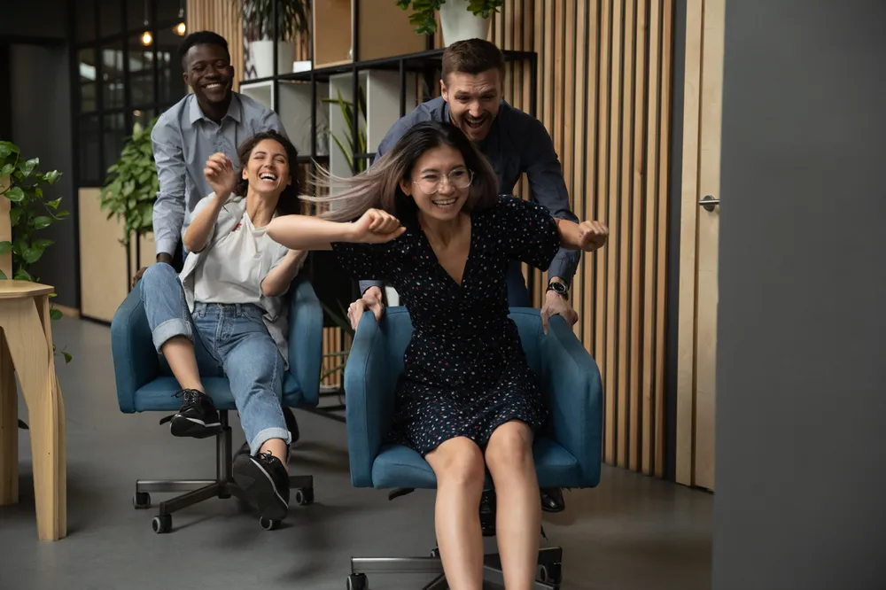 Four people in an office setting engaged in a playful activity, with two individuals sitting in rolling chairs being pushed by two others. The scene highlights a casual, fun moment with modern office furniture and wooden panelling in the background.