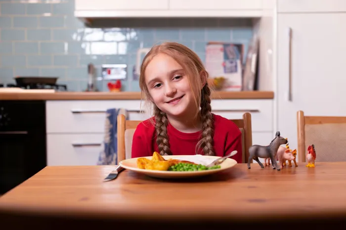 Freya smiles at the camera while sitting at a table with her dinner. Her toy animals are placed beside her on the table.