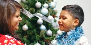 A young boy looking at a woman and smiling, wearing festive jumpers in front of a Christmas tree