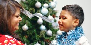 A young boy looking at a woman and smiling, wearing festive jumpers in front of a Christmas tree