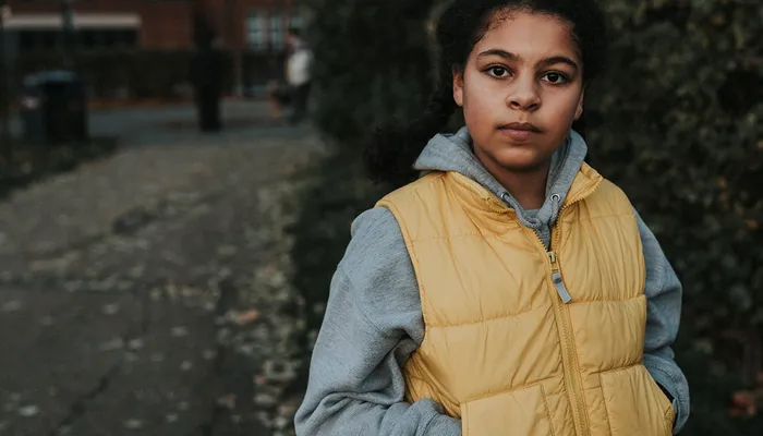 A young person wearing a yellow puffer jacket over a gray hoodie standing outdoors on a paved path with greenery and buildings in the background. The scene suggests a casual, cool-weather setting with muted natural colours and soft lighting.