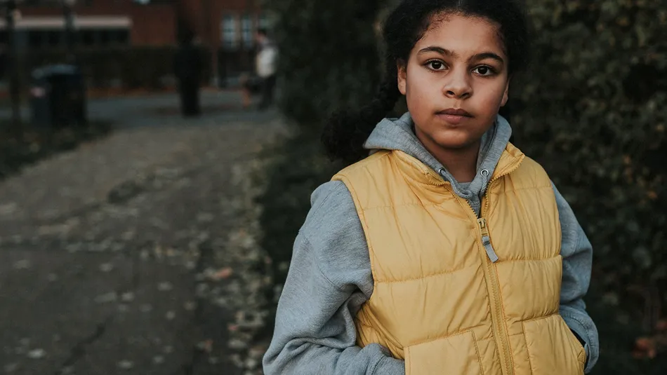 A young person wearing a yellow puffer jacket over a gray hoodie standing outdoors on a paved path with greenery and buildings in the background. The scene suggests a casual, cool-weather setting with muted natural colours and soft lighting.