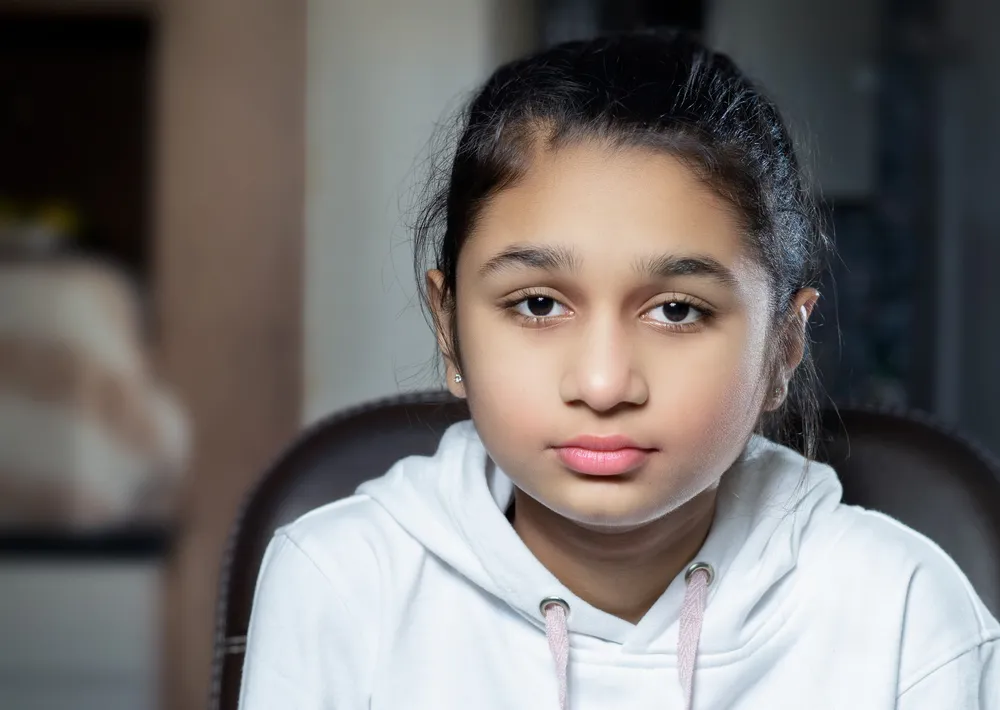 A young person wearing a white hoodie sitting indoors on a dark chair. Background includes blurred household items and soft lighting, suggesting a casual or home environment.