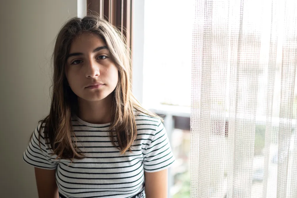 Girl standing up looking sad into camera next to curtains