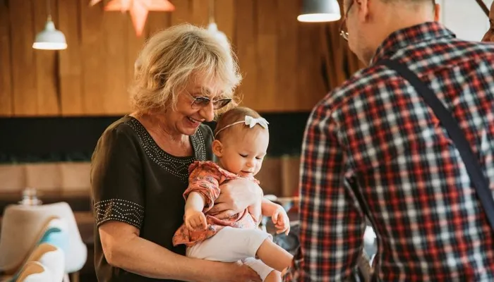 An older woman holding a baby while a man in a plaid shirt faces them in a warmly lit room with wooden walls and hanging lights. The scene captures a family interaction with cozy, casual surroundings and soft ambient lighting.
