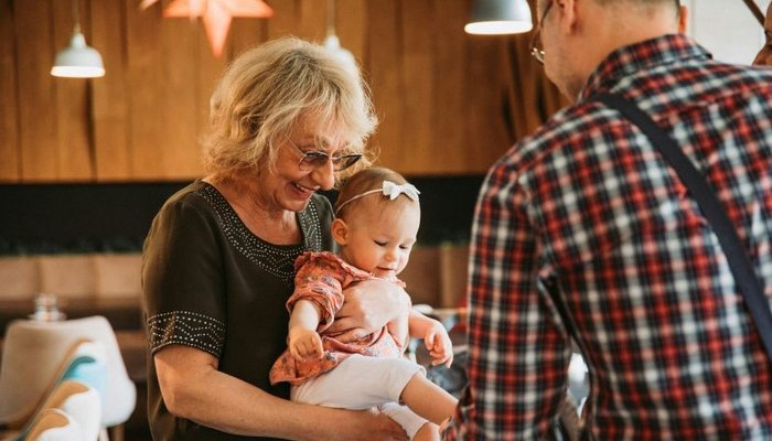 An older woman holding a baby while a man in a plaid shirt faces them in a warmly lit room with wooden walls and hanging lights. The scene captures a family interaction with cozy, casual surroundings and soft ambient lighting.
