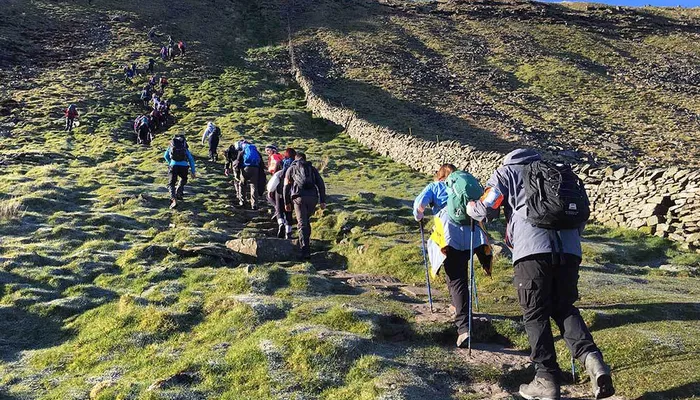 A group of hikers ascending a grassy hillside along a narrow dirt path bordered by a stone wall under a clear blue sky. The scene highlights outdoor activity with hikers wearing backpacks and using walking poles, emphasizing a challenging uphill trek