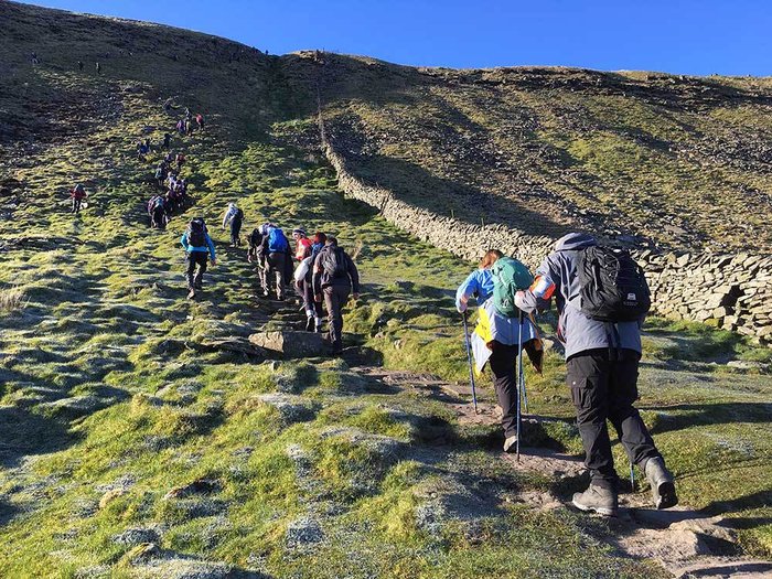 A group of hikers ascending a grassy hillside along a narrow dirt path bordered by a stone wall under a clear blue sky. The scene highlights outdoor activity with hikers wearing backpacks and using walking poles, emphasizing a challenging uphill trek