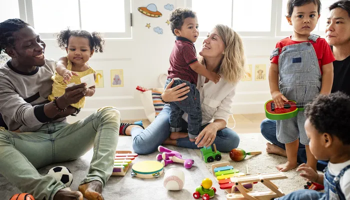 A diverse group of young children and adults playing together in a brightly lit room filled with various toys, including a xylophone, toy cars, and balls. The scene highlights interaction, learning, and play in an early childhood or daycare setting.