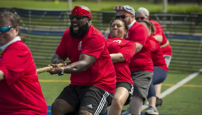 A group of people participating in a tug-of-war on a sports field, all wearing red shirts and pulling a rope with focused expressions. The scene highlights teamwork and physical effort, with individuals lined up side by side and gripping the rope tightly.