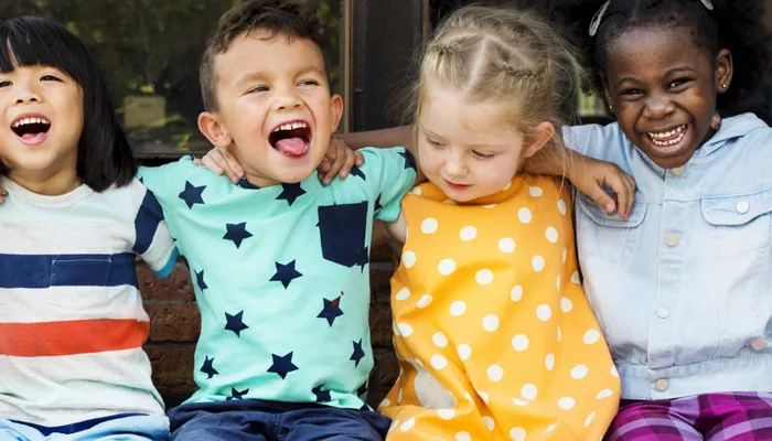 Six happy children sitting closely together on a bench outdoors, each wearing colourful clothing including patterns like stripes, stars, and polka dots. The group displays a sense of friendship and diversity, with arms around each other