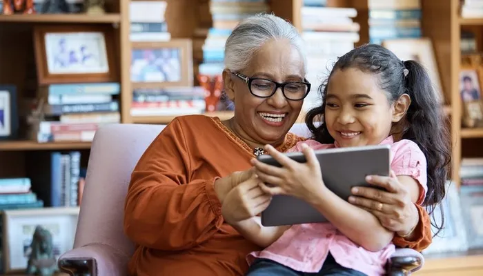 An elderly woman and a young girl sitting together in a cozy room filled with bookshelves, looking at a tablet. The scene highlights intergenerational bonding and technology use in a warm, homey environment.