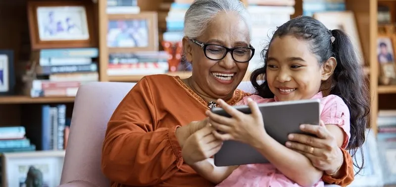 An elderly woman and a young girl sitting together in a cozy room filled with bookshelves, looking at a tablet. The scene highlights intergenerational bonding and technology use in a warm, homey environment.