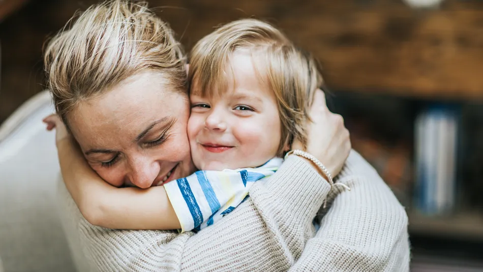 An adult and a child embracing each other closely, conveying a sense of warmth and affection. The adult wears a light-coloured knitted sweater, while the child wears a striped shirt, with a blurred background suggesting an indoor setting.