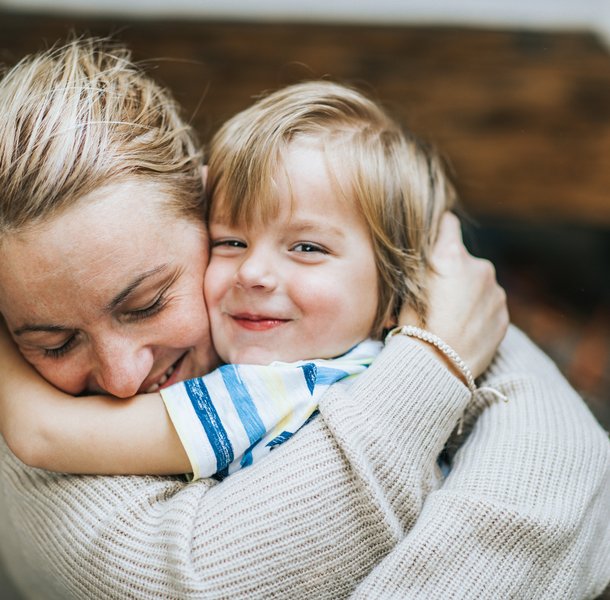 An adult and a child embracing each other closely, conveying a sense of warmth and affection. The adult wears a light-coloured knitted sweater, while the child wears a striped shirt, with a blurred background suggesting an indoor setting.