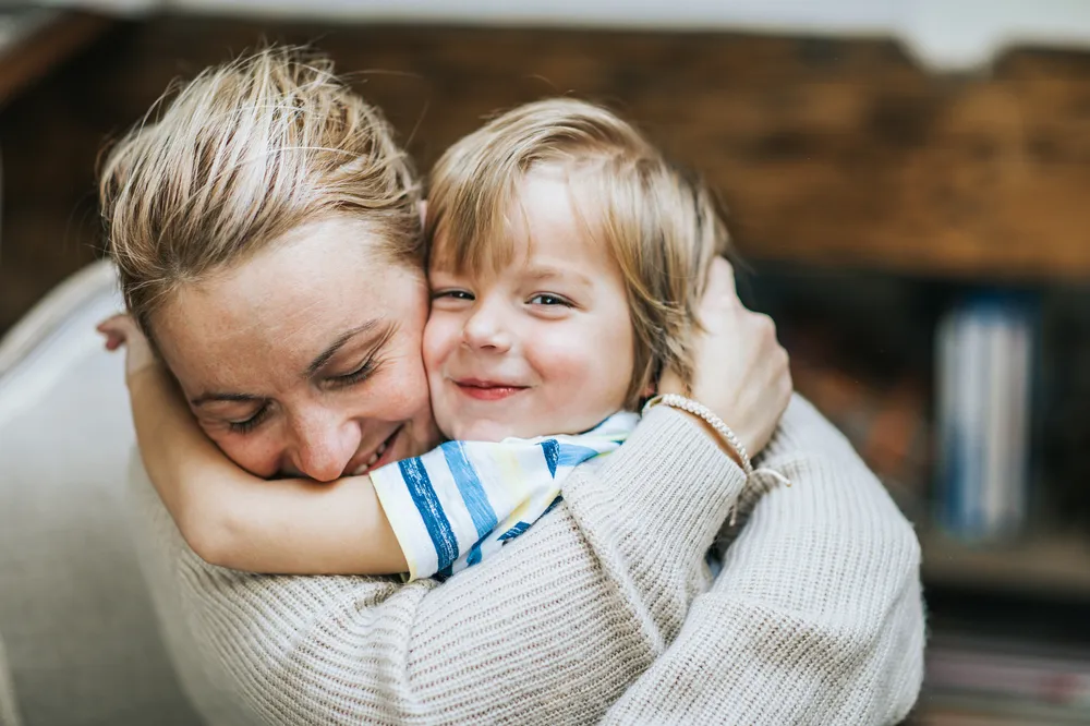An adult and a child embracing each other closely, conveying a sense of warmth and affection. The adult wears a light-coloured knitted sweater, while the child wears a striped shirt, with a blurred background suggesting an indoor setting.