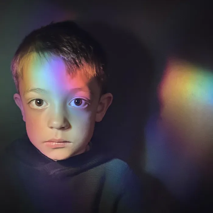 Head and shoulders of a young child against a dark background featuring a colourful rainbow light reflection on the right side. The image highlights contrast between shadowed areas and vibrant light, creating a visually striking effect.