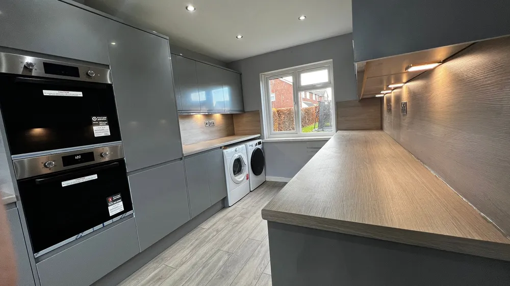 A modern kitchen with grey cabinets, two built‑in ovens, and a long countertop. Beneath the counter sit two front‑loading laundry machines, one white and one black. The room has light wood‑style flooring, and a large window showing greenery outside.