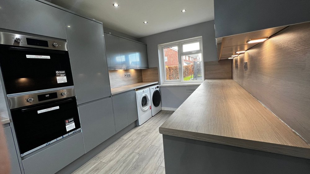 A modern kitchen with grey cabinets, two built‑in ovens, and a long countertop. Beneath the counter sit two front‑loading laundry machines, one white and one black. The room has light wood‑style flooring, and a large window showing greenery outside.