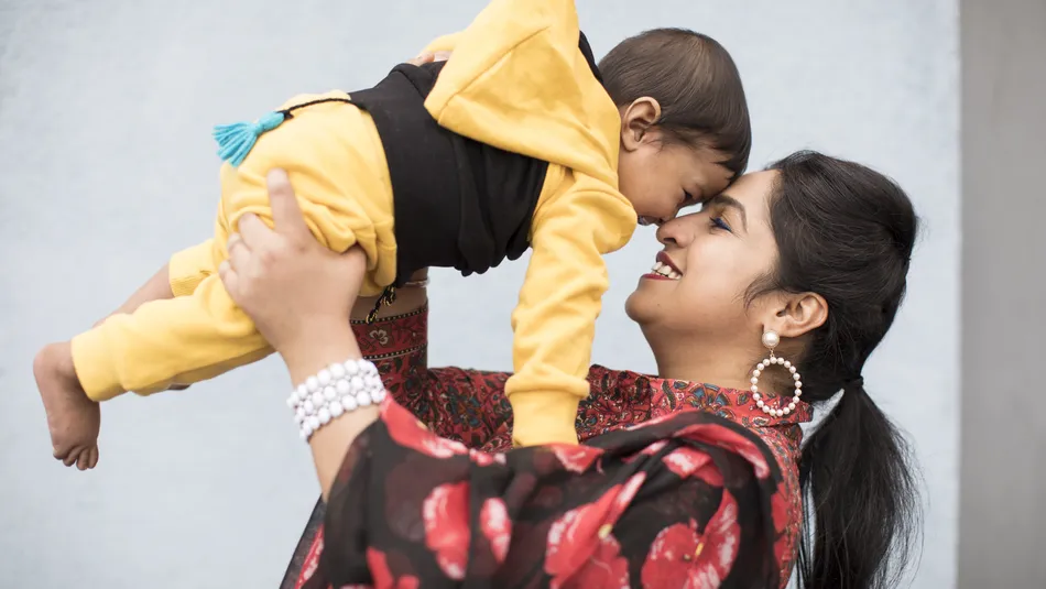 Jaspreet, wearing a black and red floral scarf, holds her baby son in a yellow and black tracksuit to her face. They smile and laugh looking at each other