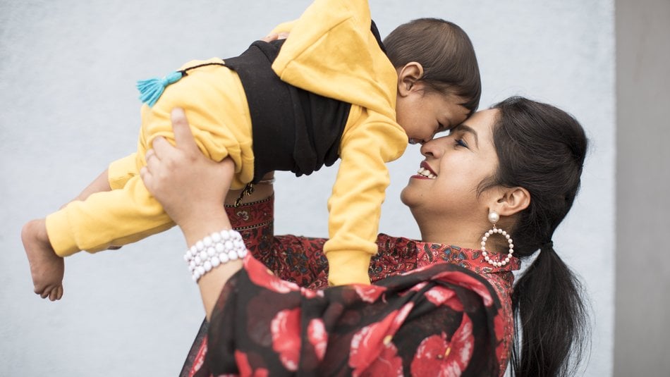 Jaspreet, wearing a black and red floral scarf, holds her baby son in a yellow and black tracksuit to her face. They smile and laugh looking at each other