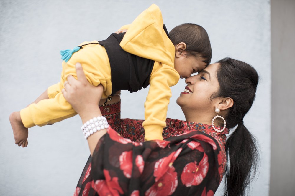 Jaspreet, wearing a black and red floral scarf, holds her baby son in a yellow and black tracksuit to her face. They smile and laugh looking at each other