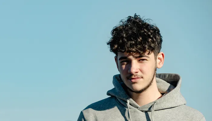 A young person wearing a light gray hoodie standing outdoors with a clear blue sky and blurred treeline in the background. The focus is on the upper body and casual attire, highlighting natural lighting and a calm outdoor setting.