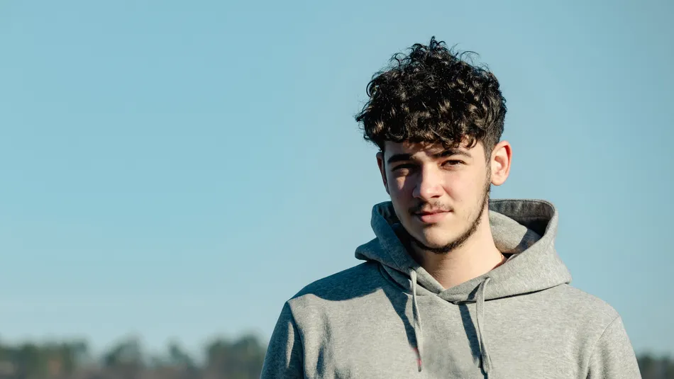 A young person wearing a light gray hoodie standing outdoors with a clear blue sky and blurred treeline in the background. The focus is on the upper body and casual attire, highlighting natural lighting and a calm outdoor setting.