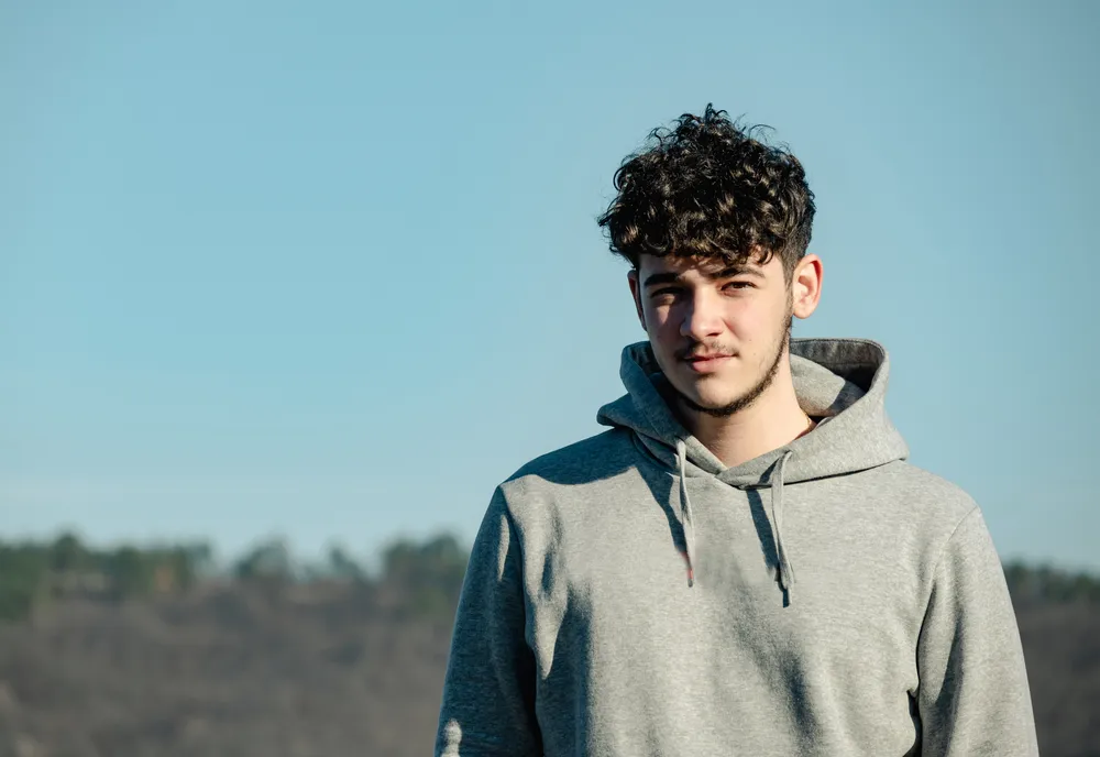 A young person wearing a light gray hoodie standing outdoors with a clear blue sky and blurred treeline in the background. The focus is on the upper body and casual attire, highlighting natural lighting and a calm outdoor setting.