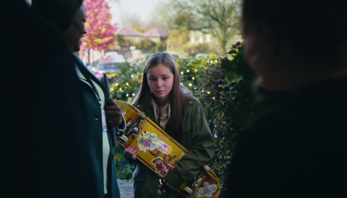 Young girl holding skateboard standing at the door look down at floor as she meets her new foster carers in John Lewis Christmas Ad