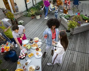 An outdoor barbecue gathering on a wooden deck with people socializing and cooking food. The scene includes a grill with meat, a table with various dishes like cupcakes, fruits, and snacks, and guests holding drinks and engaging in conversation.