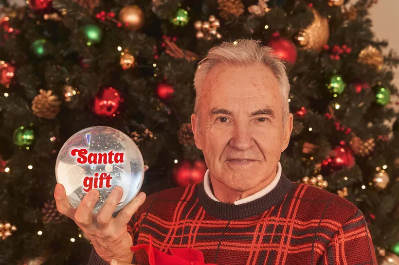 Larry Lamb sat in front of Chrsitmas tree holding a wrapped present and Snow globe that says 'Santa gift'