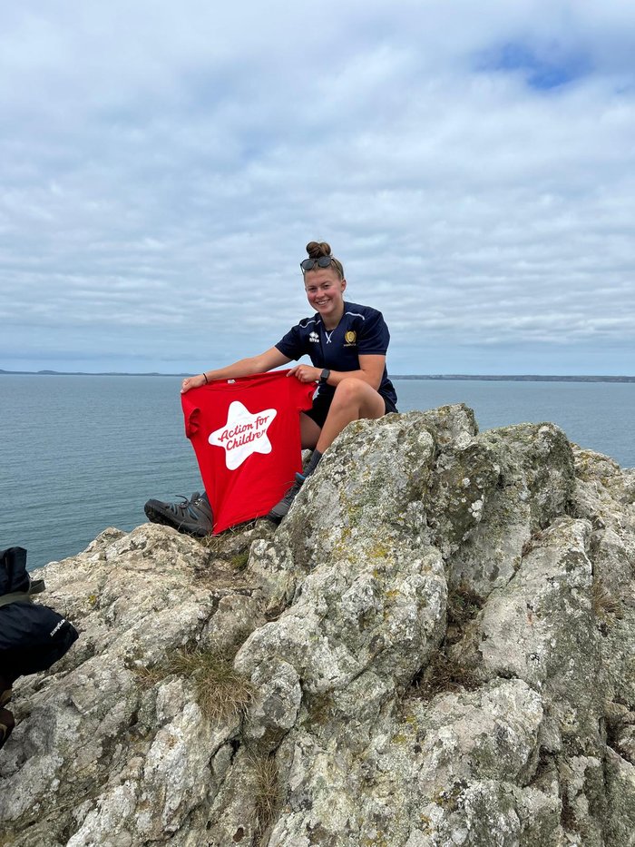 AFC fundraiser Lauren sits on top of a cliff after a challenge, holding an AFC branded tshirt