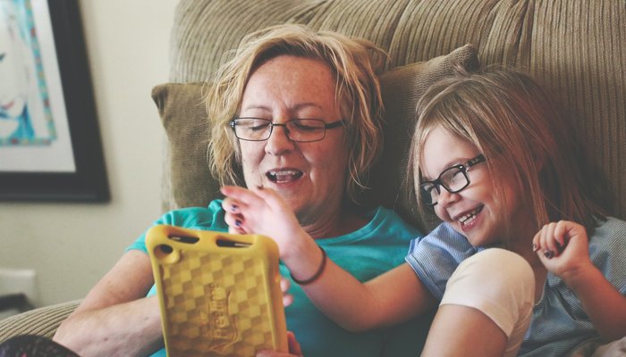 Grandmother and Granddaughter wearing glasses and sitting on the sofa together, looking at a phone and laughing