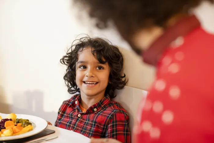 A child wearing a red checked shirt sitting at a dining table with a plate of food, facing a blurred image of an adult in a red jumper with white polka dots. The scene captures a moment of interaction, possibly during a meal or conversation.