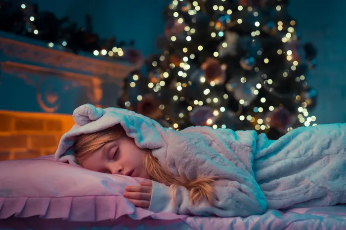 A child wearing a blue dressing gown lying on a bed with a pink pillow in front of a decorated and lit Christmas tree. The scene features warm, cozy lighting with festive ornaments and blurred lights creating a holiday atmosphere.