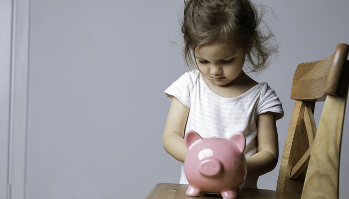 A young child standing behind a wooden chair, holding a pink piggy bank placed on the chair's seat. The setting appears to be indoors with a plain light-coloured wall in the background, emphasizing themes of saving money or financial education