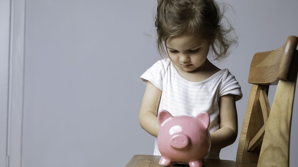 A young child standing behind a wooden chair, holding a pink piggy bank placed on the chair's seat. The setting appears to be indoors with a plain light-coloured wall in the background, emphasizing themes of saving money or financial education