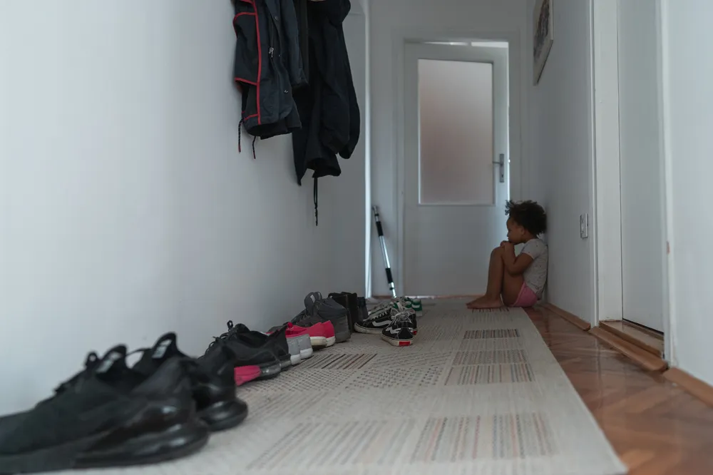 Lonely little girl sitting on the floor at the end of a long apratment corridor. Her knees to her chest and she is looking down sadly