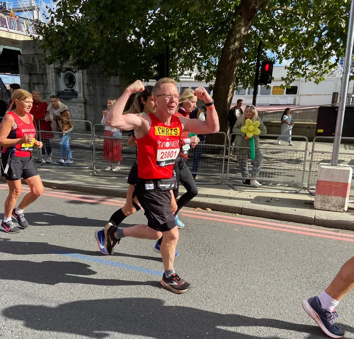Runners participating in a marathon on a city street, with one runner in a red shirt flexing his arms in celebration. Spectators stand behind metal barriers, including a person holding a large yellow flower prop, under a tree with green leaves.