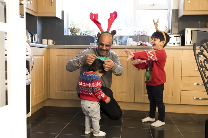 A person, wearing reindeer antlers kneeling on a kitchen floor helping a young child wear a festive headband, while another child wearing a red jumper and reindeer antlers stands nearby. Kitchen has light wood cabinets, black floor tiles and a window