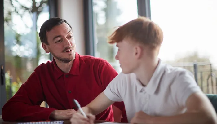 An adult and a child sitting at a table engaged in a learning or tutoring session, with notebooks and papers visible. Natural light from large windows illuminates the scene, highlighting focused interaction and educational activity.