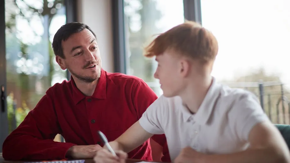 An adult and a child sitting at a table engaged in a learning or tutoring session, with notebooks and papers visible. Natural light from large windows illuminates the scene, highlighting focused interaction and educational activity.