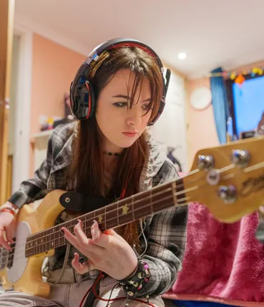 Young Carer Meghan, sitting on her bedroom floor, wearing headphones and playing her bass guitar.