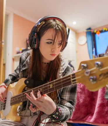 Young Carer Meghan, sitting on her bedroom floor, wearing headphones and playing her bass guitar.